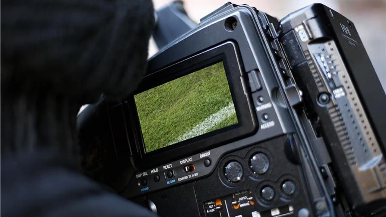 Kameramann mit Wollm&uuml;tze blickt in den Sucher einer Fernsehkamera, auf dem kleinen Monitor ist der Rasen eines Fu&szlig;ballplatzes zu sehen